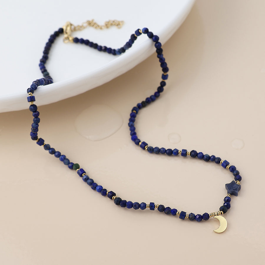 Blue beaded necklace with gold accents and gold moon charm on a beige surface