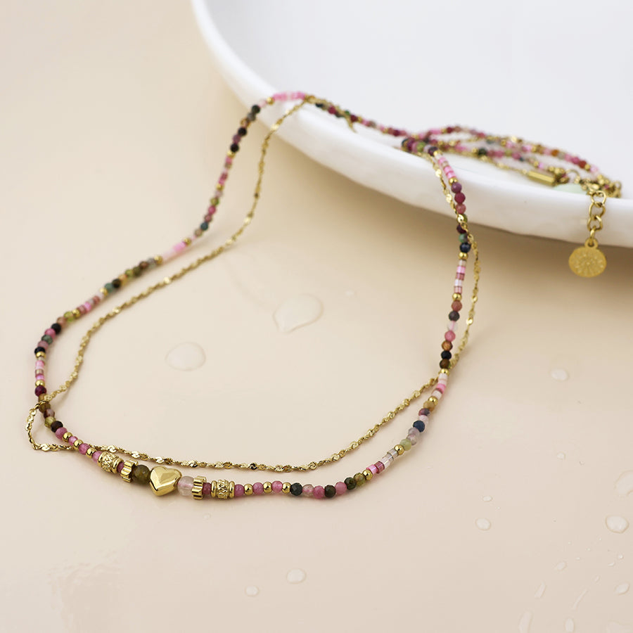 Multistrand beaded necklace with gold accents and heart charm on a beige surface. 